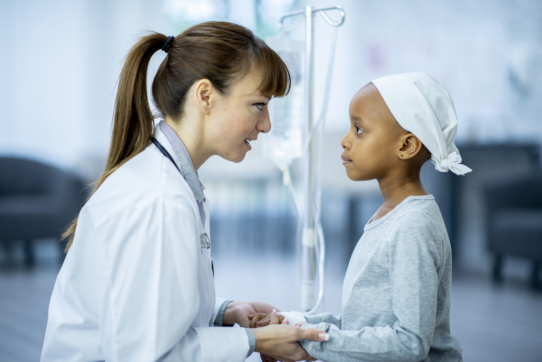 A female doctor and cancer patient are indoors in a hospital room. The doctor is holding the girl's hands to comfort her.