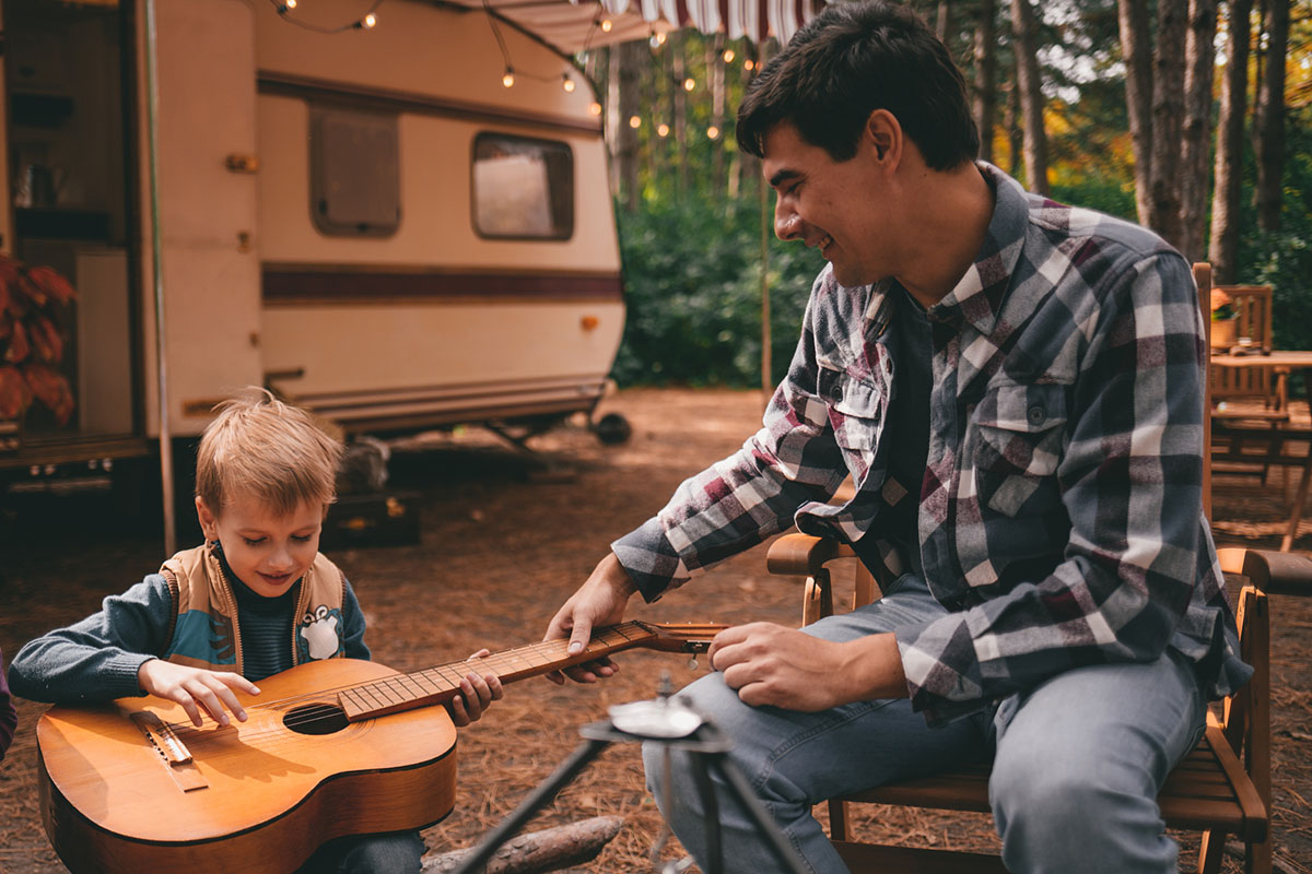 Father teaches son play guitar on camping trip relaxing in the autumn forest
