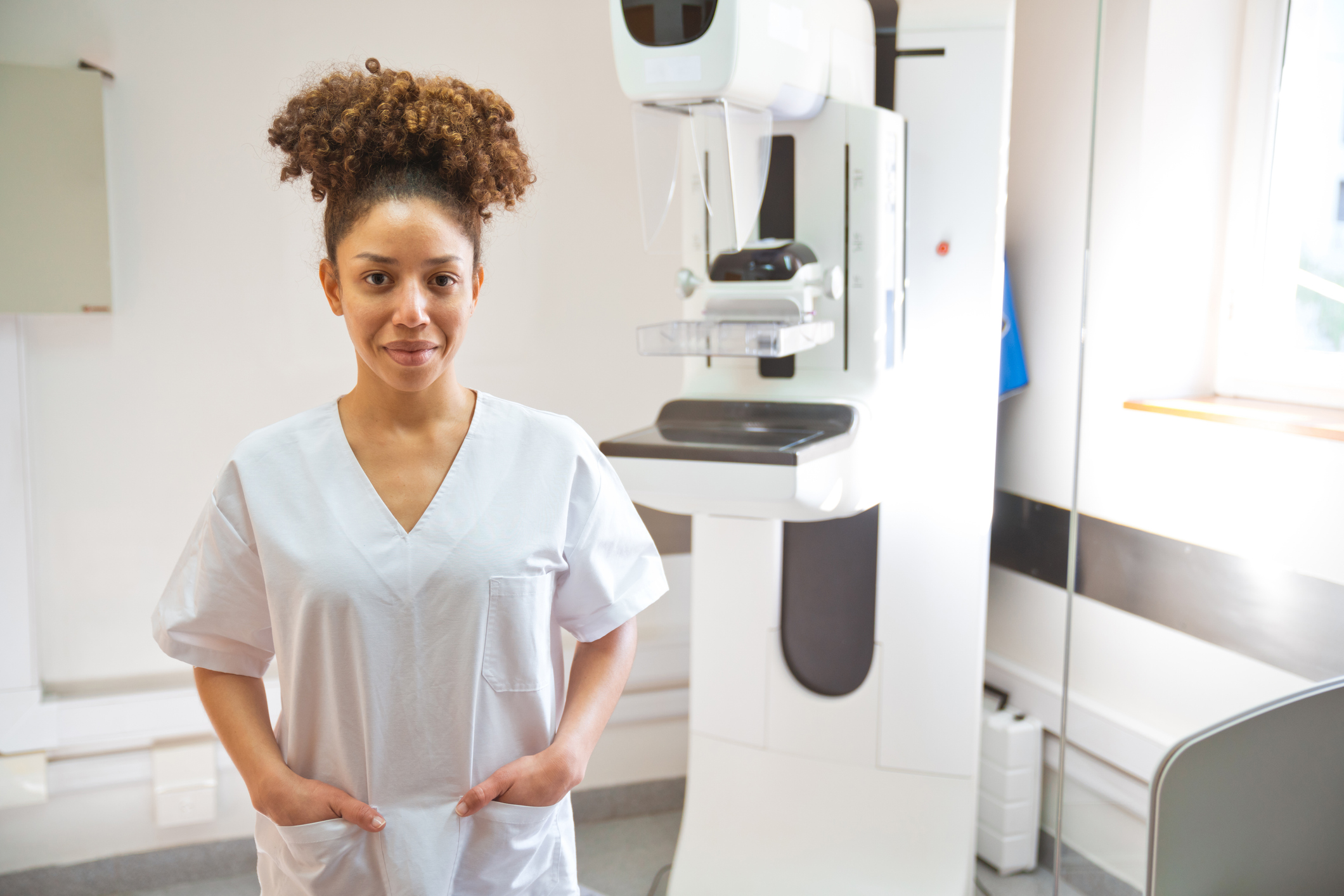 Woman standing with hands in pockets next to mammogram in medical clinic and smiling at camera
